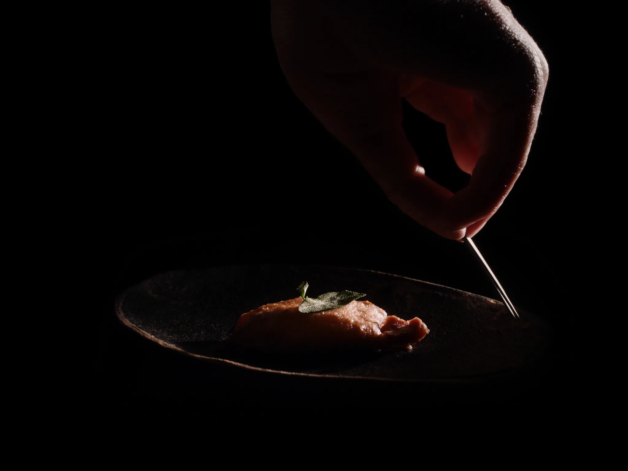 Main de chef déposant une feuille de sauge à la pince sur une assiette en grès noir