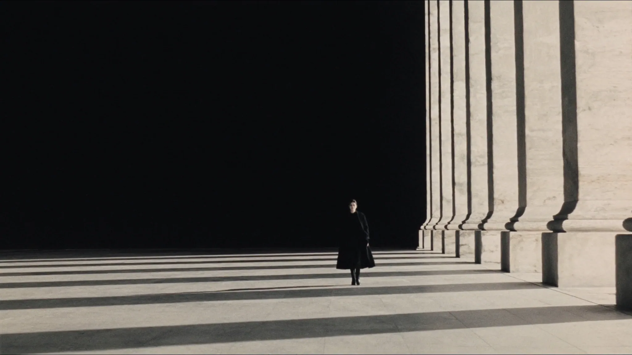 Silhouette en manteau noir marchant sous une colonnade de pierre au soleil rasant